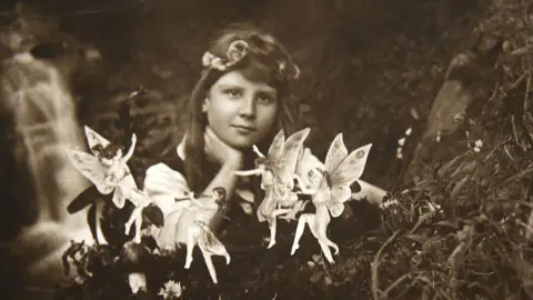 David Whittaker A black and white photo of a young girl with brown hair and a headband resting her hand on her neck and looking at the camera. In front of her are small dancing fairies. She is sitting outdoors with what appears to be a stream behind her.
