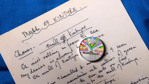 The Royal Mint A silver coin with a rainbow design and Paul McCartney's signature rests on a copy of handwritten lyrics 