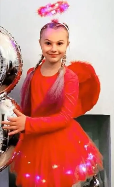Lina Savicke A family picture of Lilia wearing a red dress, halo and wings and holding a balloon