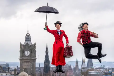 Jane Barlow/PA Stefanie Jones, dressed as Mary Poppins - in a red coat and holding a red umbrella -  and Jack Chambers, dressed as Bert -  in black trousers, red shirt and waistcoat and a black flat cap, holding a sweep - pose by jumping in the air, during a photo call on Calton Hill, with the Edinburgh skyline in the background.