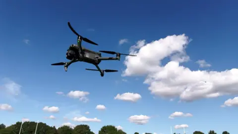 Drone flying in the air. There is a blue sky and patches of white clouds. 