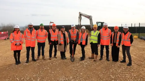 Durham County Council Eleven people standing in a line in hi-vis clothing. A man in the middle is holding a shovel. Behind them is a large digger and the brown earth they are standing on is flat and covered in small partially buried rocks.