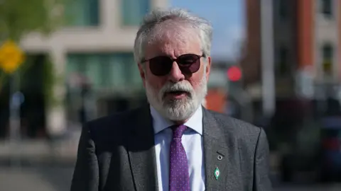 Gerry Adams, with grey hair and a grey beard, wearing sunglasses, a grey suit, light blue shirt and purple tie. The background of the image is blurred. 