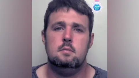 A mugshot of a man wit short black hair, a beard and a grey or black top.