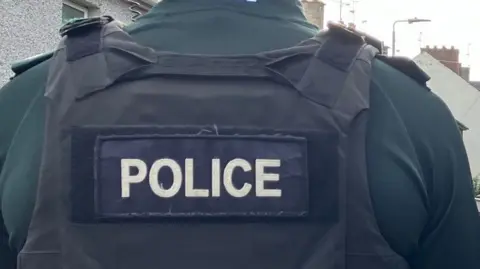 A close-up of a PSNI officer's uniform from behind.  The man is wearing a dark green PSNI polo shirt under a black protective vest which shows the word "police" in large white lettering.