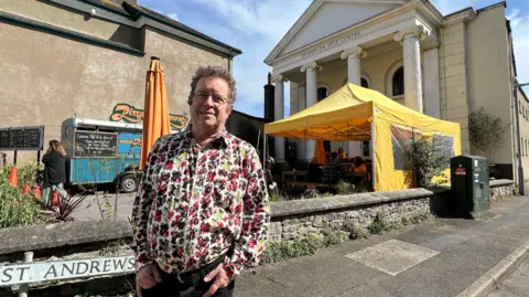 The picture shows a sunny street scene outside a historic-looking building with tall white columns. A bright yellow canopy is set up in front of the building, and people appear to be sitting underneath it, as if at a small market, café space or community event. In the foreground, a person is standing on the pavement wearing a colourful floral shirt and dark trousers, with their hands in their pockets.