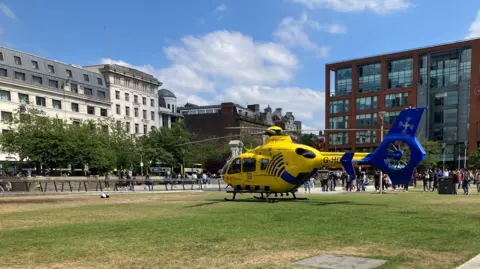 The yelow and blue air ambulance in the middle of Piccadilly Gardens, on the grass