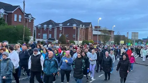 People walking in west Belfast in various items of clothing. There are brown brick buildings to the left and street lights in the distance.