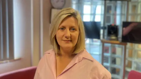 A blonde hair woman wears a pink shirt. Behind her left shoulder are two black tv screen mounted on a wall. Below the TV's are tow red sofas. Over her right shoulder is a closed white window blind.