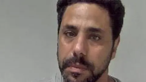 West Mercia Police A man with short black hair and a short black beard is pictured in a grey sweatshirt in front of a neutral-coloured background.