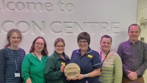 Myeloma UK Six people with a circular wooden award, which reads Myeloma UK Clinical Service Excellence Programme.