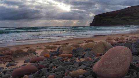 Iain Kelman Rocky beach with smooth, rounded stones scattered across the sand. Small waves roll in from the sea, and sunlight breaks through clouds over distant coastal cliffs.
