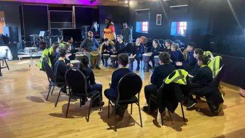 A group of primary school children sitting on chairs in a circle, there are 3 men at the top of the circle teaching music. One is sitting down playing a drum while the other two men are standing 