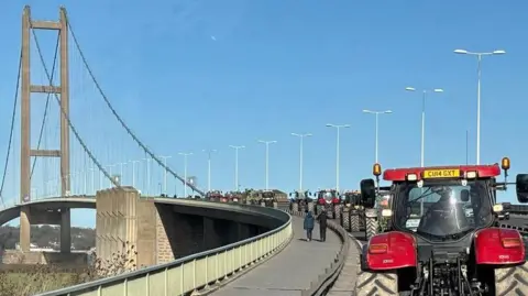 A row of tractors make their way over the Humber Bridge.  The bridge curves away to the left and the tractors disappear around the curve.  It is a sunny day with bright blue sky.