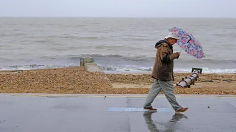 Stephen Squirrell A man wearing a white hat, brown coat and brown shoes walks along a seafront during a rainy and cloudy day. He is holding a multi-coloured umbrella over himself. 
