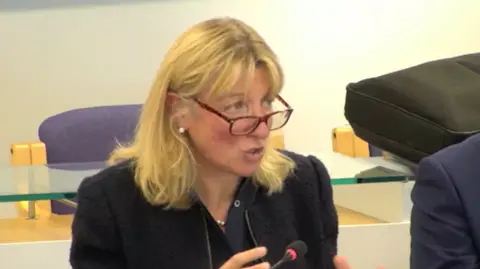 East Sussex County Council Jo Shiner has blond hair and glasses and is wearing a black suit. She is sitting down and speaking, answering questions from a panel of councillors in a council chamber