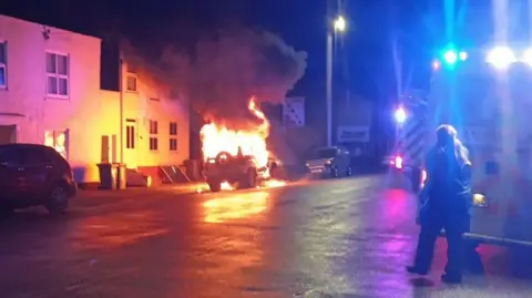The rear of a 4x4 car can be seen beneath a large quantity of flame, with thick grey smoke above on a road in Wisbech. It is lighting up the walls of the white buildings to its left. On the right, is the back of emergency services, including an ambulance, and a person walking towards the incident. 