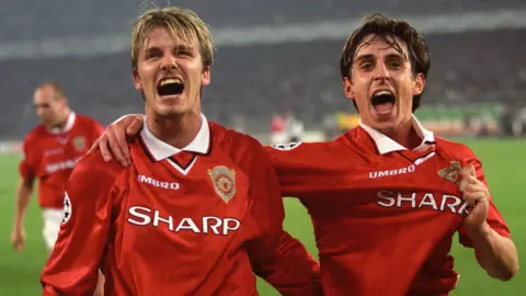 PA Media Manchester United's David Beckham and Gary Neville celebrate at the end of the game in 1999, both have hugely emotive facial expression and are wearing the red and white kit of the club they played for. A number of other players can be seen on the pitch which is seen in the background