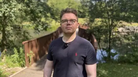 Conservative leader Councillor Anthony Mullen standing in front of the pond at Barnes Park. He is wearing a black T-shirt and glasses. His short brown hair is swept back.