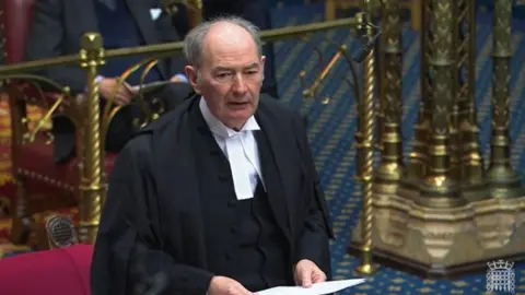 Lord Forsyth dressed in a black cloak and white shirt. He is holding a piece of paper as he addresses the House of Lords.