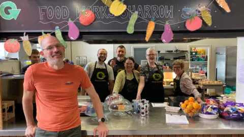 A man in an orange T-shirt ad green trousers stands in front of a group of people in a kitchen. The kitchen is adorned with paper cut outs of vegetables. On the counter tops is fruit as well as some cakes.