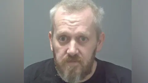 Suffolk Constabulary Mugshot of Peter Nash. It shows him with a brown and white beard. He has light grey hair and is wearing a dark navy t-shirt. 