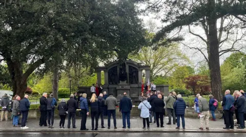 People stood in a crowd facing the Titanic Engineers' Memorial in East Park the memorial has an angel stood tall in the centre and she is flanked by workers.