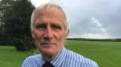 Peter Rogers wearing a stripy blue and white shirt and a dark tie, with a field of grass in the background and a tree to the left.