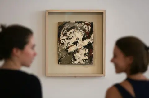 Getty Images Sotheby's employees pose for a picture in front of an artwork owned by the David Bowie, Head of Gerda Boehm by Frank Auerbach, painted in 1965. It is an abstract-looking artwork, primarily white and dark colours. 