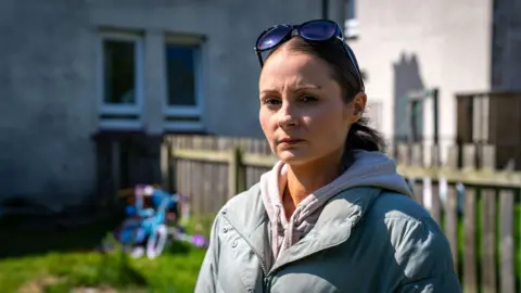 Laura, in a grey puffer jacket and grey hoodie, is standing in a garden. A child's bike can be seen behind her.