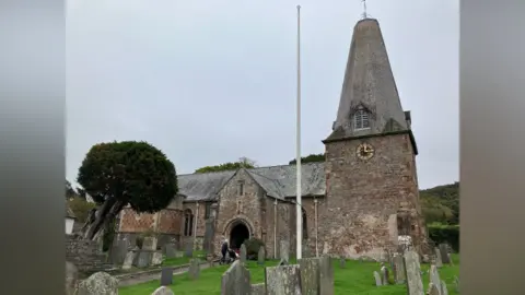 Freeline Gfx The church stands in the background, with its spire rising up through the image. The church yard is in the foreground, with a person walking through the door to the church. The sky is grey.