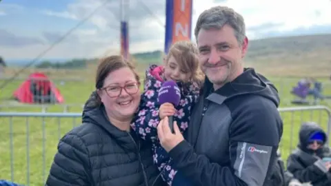 Nadia and Pete holding Amber, aged 4, in his arms. They are all smiling at the camera.