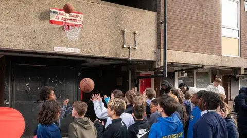 PinPep Children play with a basketball in front of a No Ball Games sign that has been transformed with a basketball hoop. 