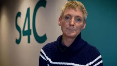 Sian Doyle poses against wall which is painted white and blue and has S4C written on it. She wears a navy jumper with white stripes and has short, blonde hair.