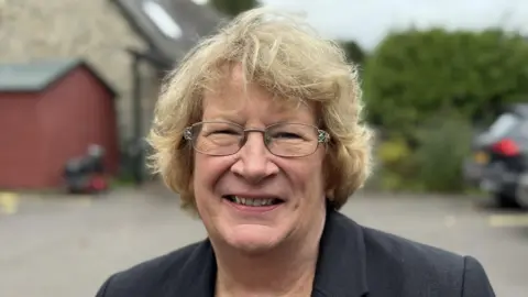 A woman with short dark blonde hair and silver glasses. She is wearing a black suit jacket and is standing in a car park with a parked car in the backgound near a brown brick building.