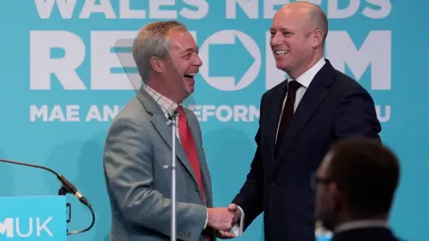PA Media Nigel Farage shaking hands with Dan Thomas on stage at a rally in Newport. The light blue set behind them says Wales needs reform in white lettering.
