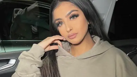 Other Woman with long brown hair in a car wearing a hoodie.
