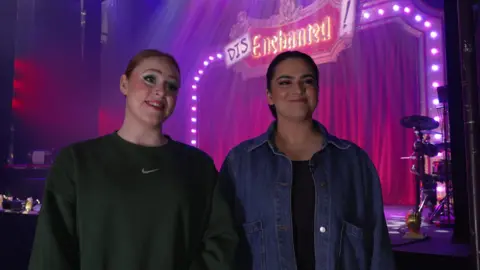 A woman with auburn hair tied back in a dark green sweater, next to a woman with dark brown hair tied back, in a black t-shirt and blue denim jacket. Behind them is a stage lit up in purple lighting. There is a red curtain and a sign reads "Disenchanted!"