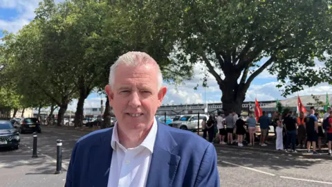 John Watson standing in front of a picket line wearing a suit and a shirt