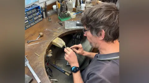 Humphrey and Hawkins A man sharpening a golden dart in a workshop.