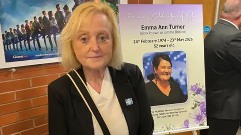 Deborah Montague. She has short blonde hair and is stood in front of a sign for Emma Britton's celebration of life. Deborah is looking at the camera with a smile on her face.