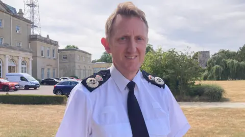 A man is standing outside a police building - he is wearing a white shirt and a black tie. The shirt is police uniform, with decorative black pads on the top of either shoulder section.