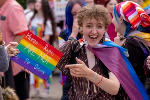 PA Media A Pride parade participant draped in a purple, blue and pink flag holds a rainbow-coloured sign that reads “Human Rights Are Not Up For Debate.” 