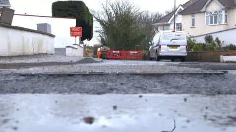 A wide shot of a road closed for resurfacing. There is a van parked on one side of the road, and a large red 'road closed' sign.