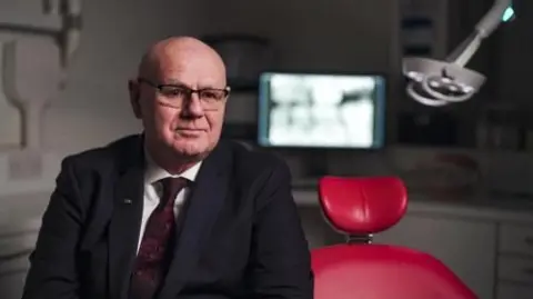 A bald man in glasses and a suit sits on a red dentist's chair in a surgery. There is a blurred computer screen in the background, showing what looks like an x-ray of teeth.  
