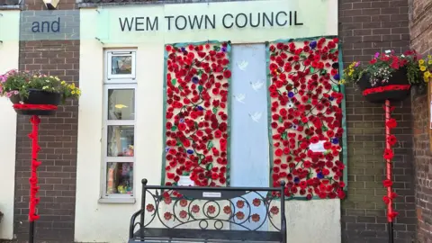 Wem Town Council A black memorial bench with metal poppies on it. On a building behind it are cascades of red knitted and crocheted poppies hanging down. On two tall flower planters on either side of the bench, poppies and red threads wind around the poles