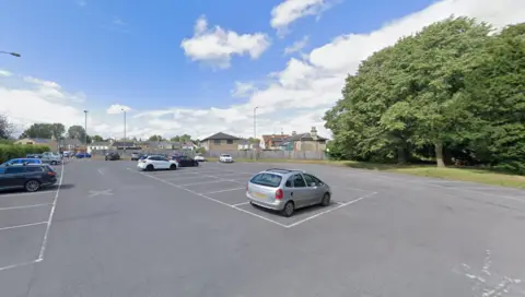 Google Maps A car park with several cars parked in it and trees behind it. There are many empty bays.