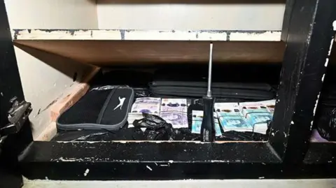 West Yorkshire Police A hidden compartment at the bottom of a black wardrobe propped open by a screwdriver. Inside, a sports bag, black suitcase and black bin bags with wads of £20 and £5 notes can be seen.
