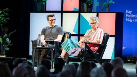 Adam Tatton-Reid/Hay Festival A man and a woman sat on a stage with crowds in front of them. There is a small table with water in front of them. The man has short brown hair and is wearing a grey t shirt and glasses. The woman is wearing a multicoloured stripy dress and glasses and has short grey hair. Behind them is a multicoloured background, plants and the Hay festival logo.