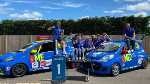 The motorsports team from West Suffolk College gather around their two race cars and trophy haul. The team are dressed in blue polo shirts and two team members sit on top of the cars.